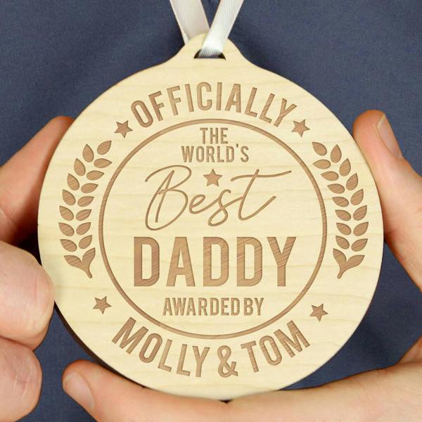 A close-up of a round wooden medal engraved with "OFFICIALLY THE WORLD'S Best DADDY AWARDED BY MOLLY & TOM" being held by two hands.