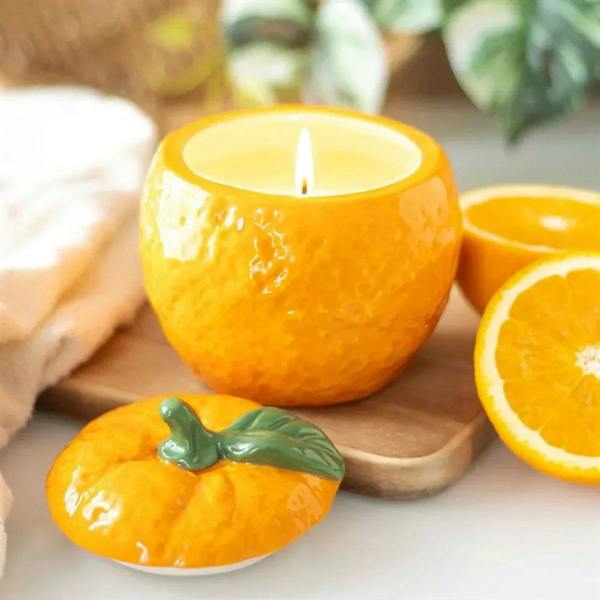 Orange-shaped candle jar with lit wick, textured ceramic, green leaf on lid