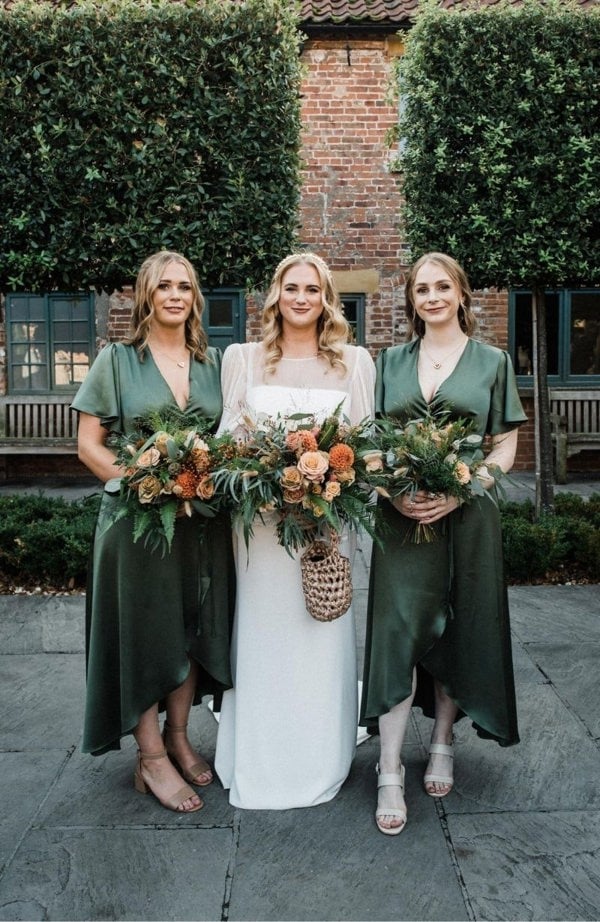 Bride with two bridesmaids in green dresses holding floral bouquets outdoors.