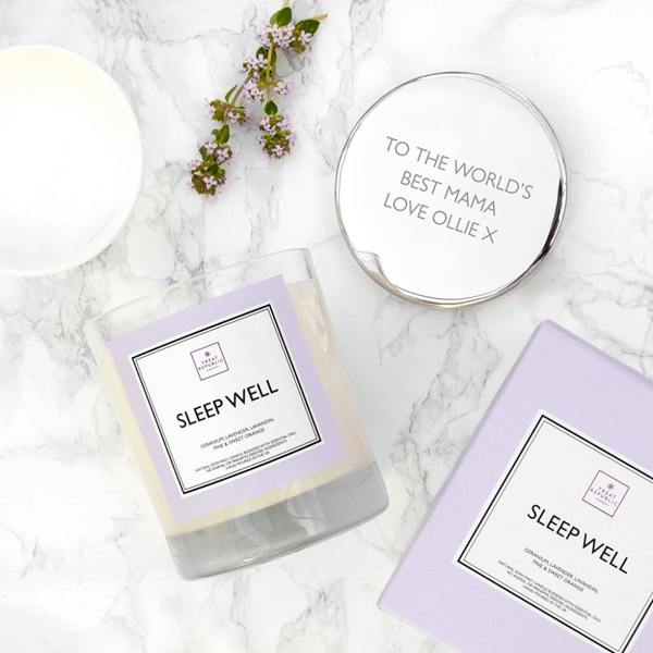 An overhead flat lay view of a clear glass 'Sleep Well' scented candle tumbler with a pastel purple label on a white marble surface. Beside it sits its polished silver metallic lid engraved with the text 'TO THE WORLD'S BEST MAMA LOVE OLLIE X'. A matching lilac gift box, a white ceramic dish, and a small sprig of purple flowers complete the styling.
