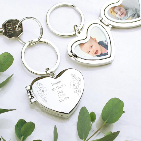 A silver heart-shaped locket keyring shown in three states: closed, partially open, and fully open. The closed locket features an engraved floral design with the text "Happy Mother’s Day Love Amelia." When opened, the locket reveals two heart-shaped photo inserts featuring high-quality portraits of smiling babies. The keyrings are displayed on a white marble surface accented with green eucalyptus leaves.