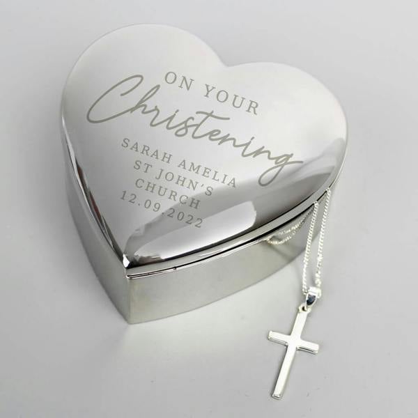 A polished, heart-shaped nickel trinket box engraved with "On Your Christening" and personalised details, with a silver-plated cross necklace draped over the side.