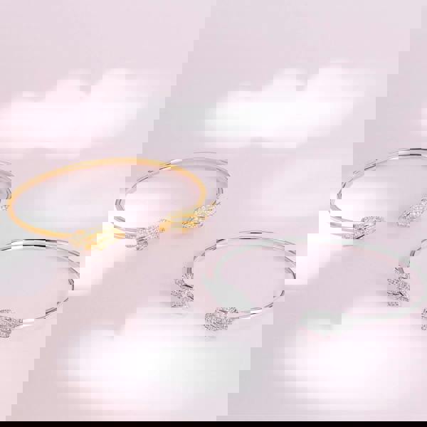 Two gold and silver bangles with feather designs on a pink background. Flexible open-wing sterling silver bangle by BLOOMTINE, set with 98 ethically sourced lab moissanites. This tarnish-resistant, hypoallergenic bracelet features a mirror-like rhodium finish and an innovative clasp-free fit, making it a premium milestone gift for 'Extraordinary Humans