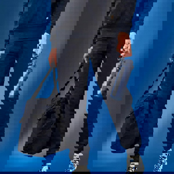 Person holding a dark bag and a blue water bottle against a blue background wearing black sweatpants and sneakers