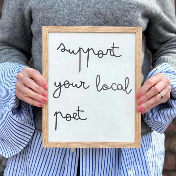 A woman in a grey wool jumper and blue striped shirt holding a solid oak framed white cotton typography print displaying the lowercase handwritten-style quote "support your local poet".