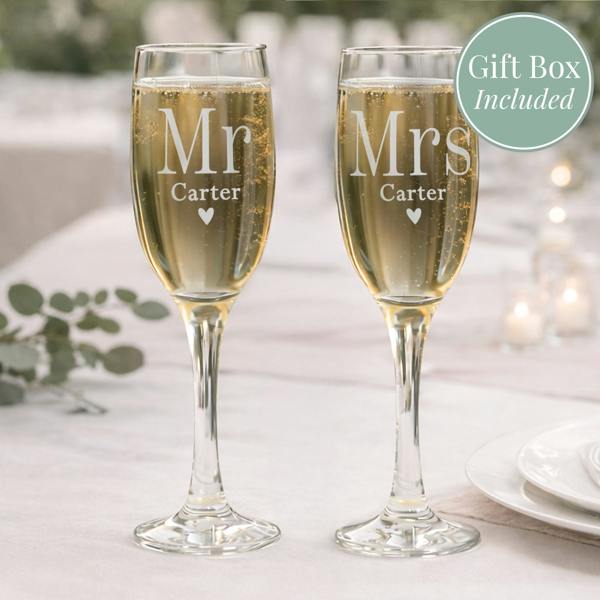 A matching pair of personalised Mr & Mrs wedding champagne flutes by Abode Home & Gifts, engraved with the custom family surname "Carter" above a small heart, filled with bubbly wine on a wedding reception table. A green circular badge reads "Gift Box Included".