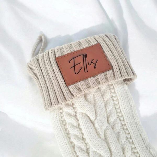 Close-up of a personalised cream cable-knit Christmas stocking cuff with an engraved faux leather name patch.