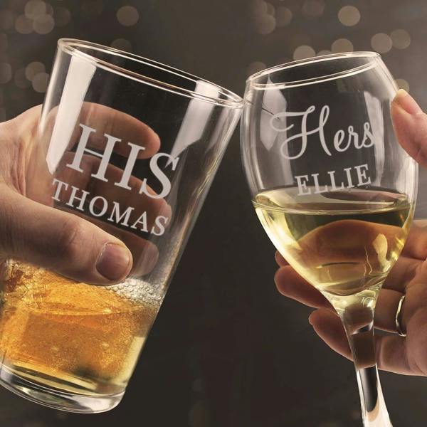 A close-up lifestyle shot of a couple clinking a matching glass set by Abode Home & Gifts in a celebratory toast. A hand holds a pint glass filled with golden lager engraved with "HIS THOMAS", and another hand holds a wine glass filled with white wine engraved with "HERS ELLIE".