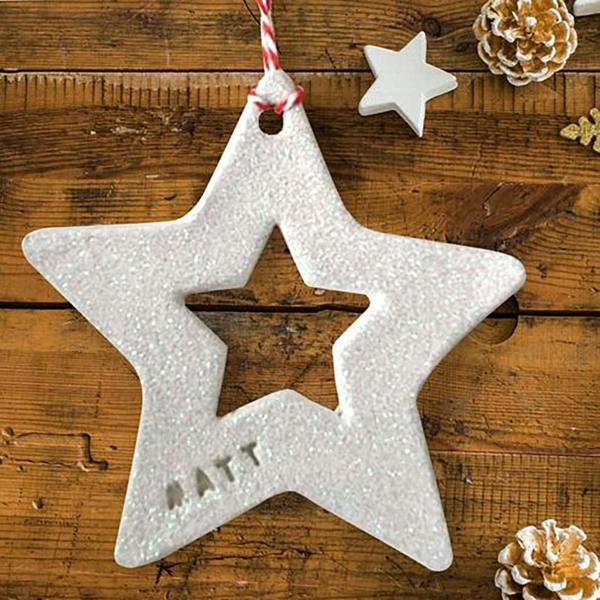 A handcrafted white clay star Christmas tree decoration hand-stamped with the name 'MATT' and coated in iridescent white glitter, hanging from a red and white twine loop over a wooden surface.