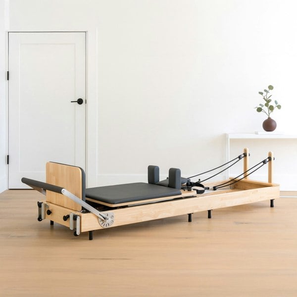 Pilates reformer machine in oak wood with a black padded platform, adjustable straps, and a minimalist design, set in a bright room