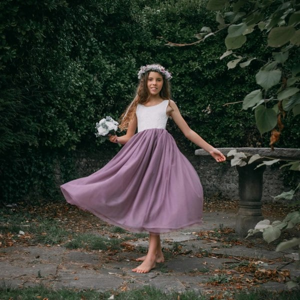 Young girl in a purple skirt and floral headband standing outdoors with greenery around.