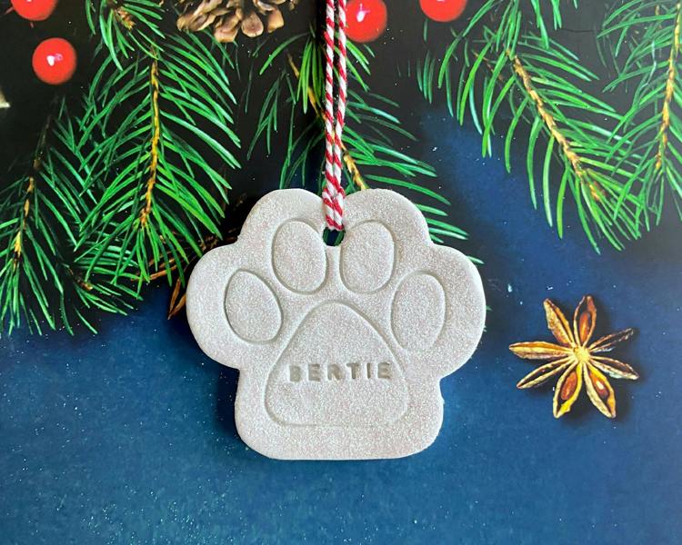 A handcrafted white clay paw print Christmas decoration hand-stamped with the name BERTIE, featuring a red and white hanging twine loop against a festive pine tree background.