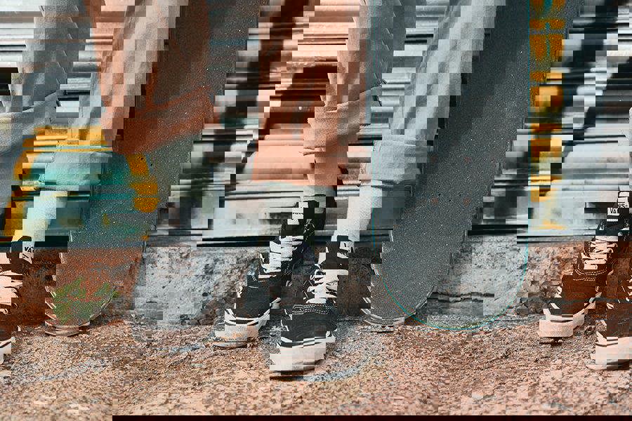 Person wearing black Vans shoes with white soles and mint green socks, standing next to a skateboard on a street.