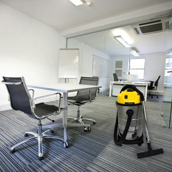 MAXBLAST 30L Industrial Vacuum Cleaner with a yellow top, stainless steel body, and black hose, positioned in a modern office setting