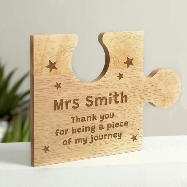 A natural wooden jigsaw piece engraved with stars, the name Mrs Smith, and the message Thank you for being a piece of my journey standing upright.