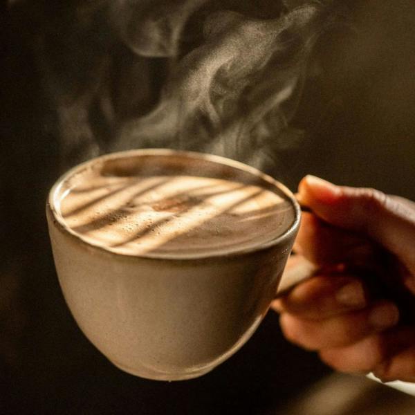 A hand holding a ceramic mug of frothy, steaming hot drinking chocolate illuminated by dramatic shafts of sunlight against a dark background.