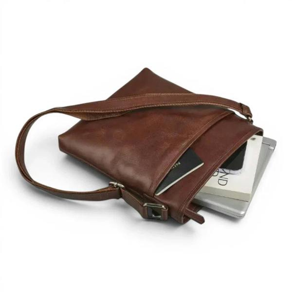 A cognac leather crossbody bag with a long strap, shown open with a passport and books inside, on a white background.