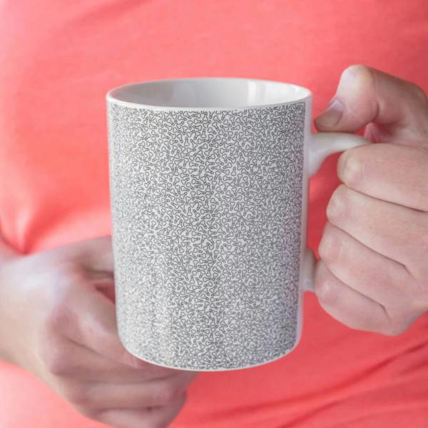A person holding a white ceramic mini mug covered in a fine, detailed pattern of tiny cheeky illustrations against a coral pink top.