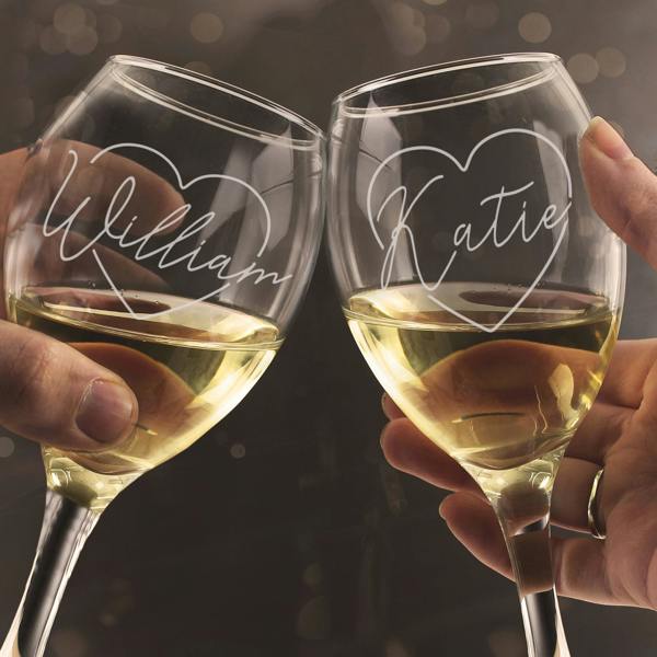 Two hands clinking a pair of personalised wine glasses by Abode Home & Gifts in a celebratory toast, filled with white wine, showing the custom script names "William" and "Katie" inside an etched heart outline against a warm, blurred background.