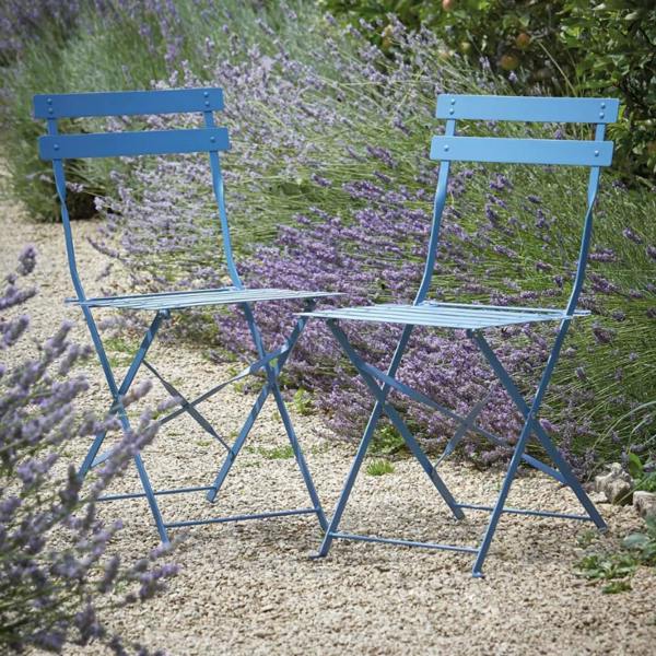 A matching pair of ocean cove blue metal folding bistro chairs styled on a gravel path next to blooming purple lavender bushes.