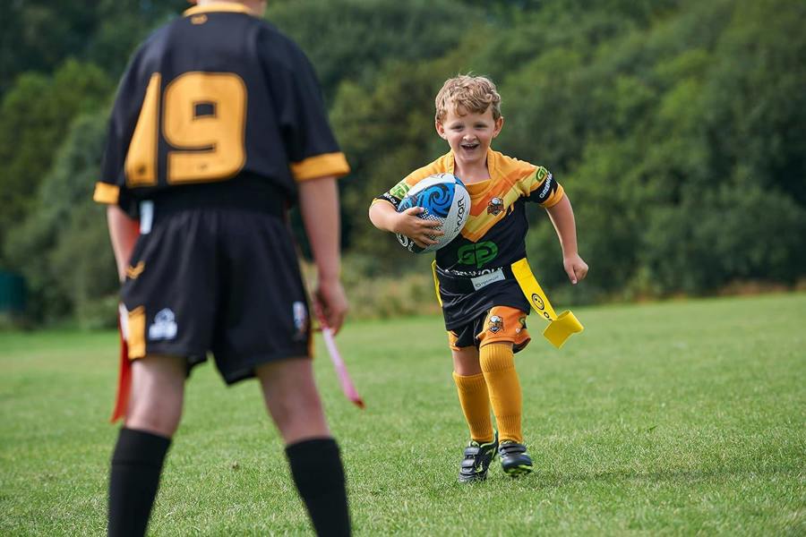Young player running with ball in yellow Optimum tag belt toward a defender in red flags, on grass