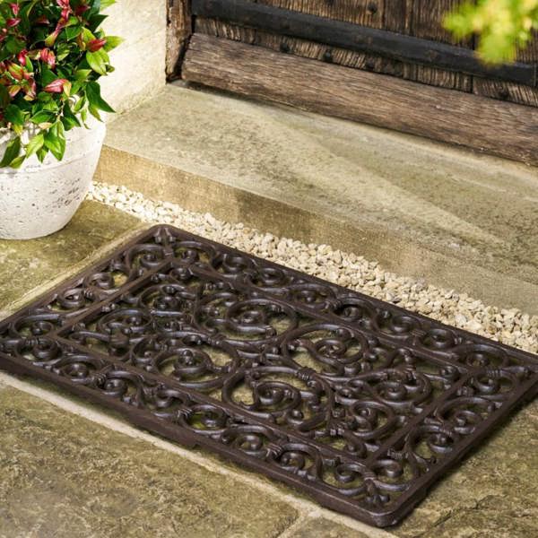 An ornate dark brown cast iron doormat placed on stone steps outside a rustic wooden door by Abode Home & Gifts.