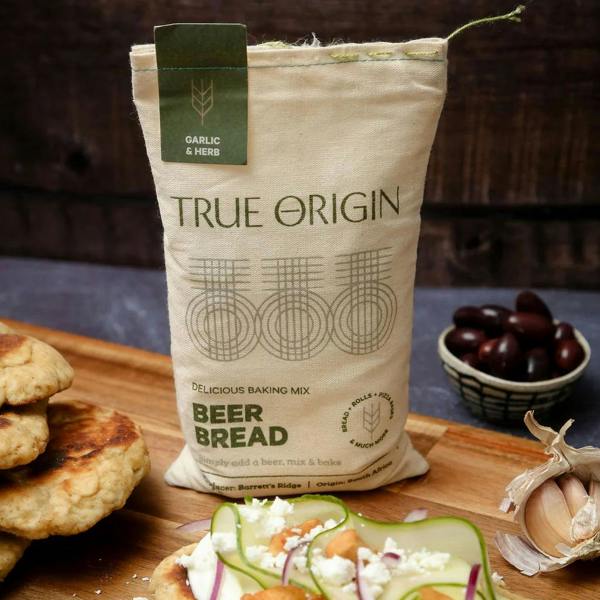 A fabric sack of Garlic and Herb beer bread mix standing behind a freshly made flatbread topped with cream cheese, cucumber ribbons, and feta on a wooden board.