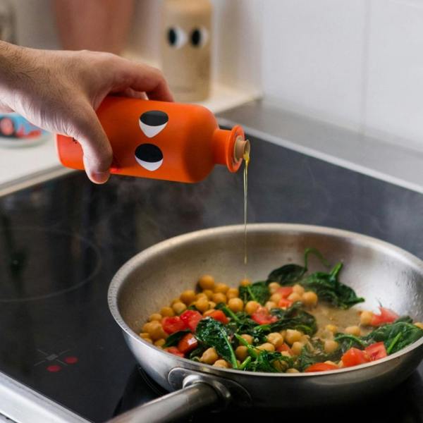 Person pouring chilli infused extra virgin olive oil from the orange bottle into a frying pan with chickpeas, spinach, and tomatoes.