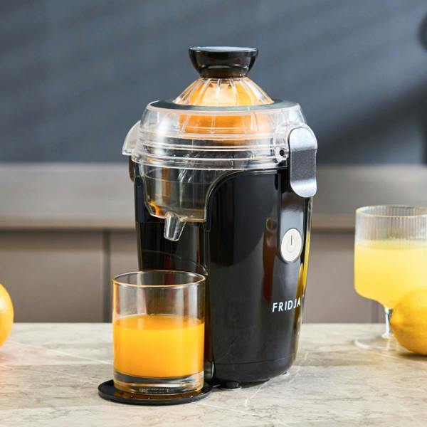 A Fridja f800 Automatic Citrus Juicer in black sits on a countertop with a halved orange on top, dispensing fresh juice into a glass. Nearby are another filled glass and an orange; the Fridja brand name is visible on the juicer.