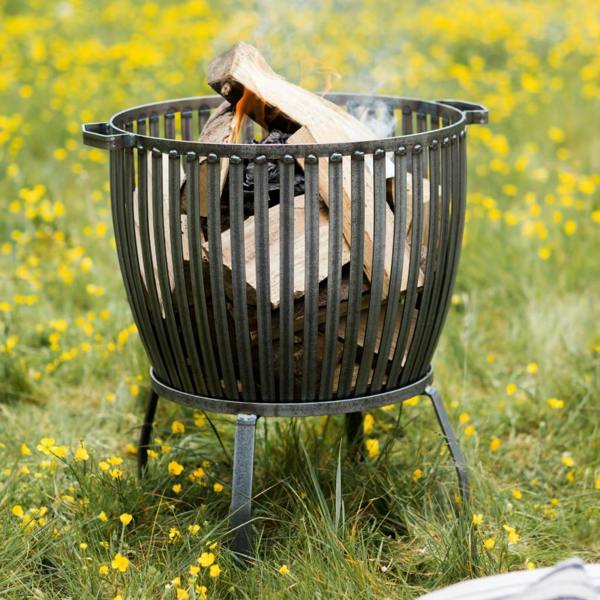 The large Barrington Fire Pit in raw metal, shown with a roaring log fire in a flowering meadow garden setting.