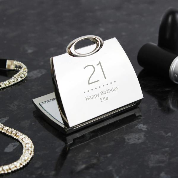 A high-angle studio shot of a shiny, silver nickel-plated handbag-shaped compact mirror, open on a dark marble surface. The lid is engraved with the number "21" and the message "Happy Birthday Ella." A sparkling rhinestone bracelet sits to the left, and a tube of lipstick is positioned to the right.
