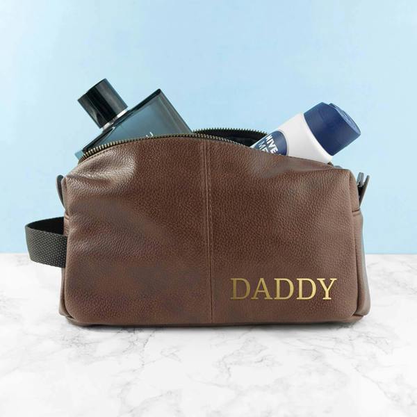 A brown grained NuHide leather wash bag personalised with Daddy in gold lettering, filled with men's grooming products.