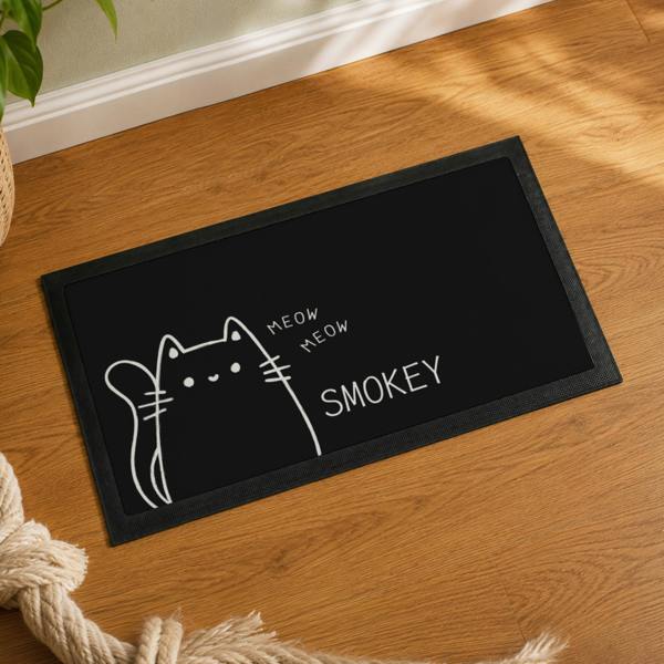 High-angle shot of a black personalised cat bowl mat featuring a cartoon cat illustration and the name SMOKEY on wooden flooring.