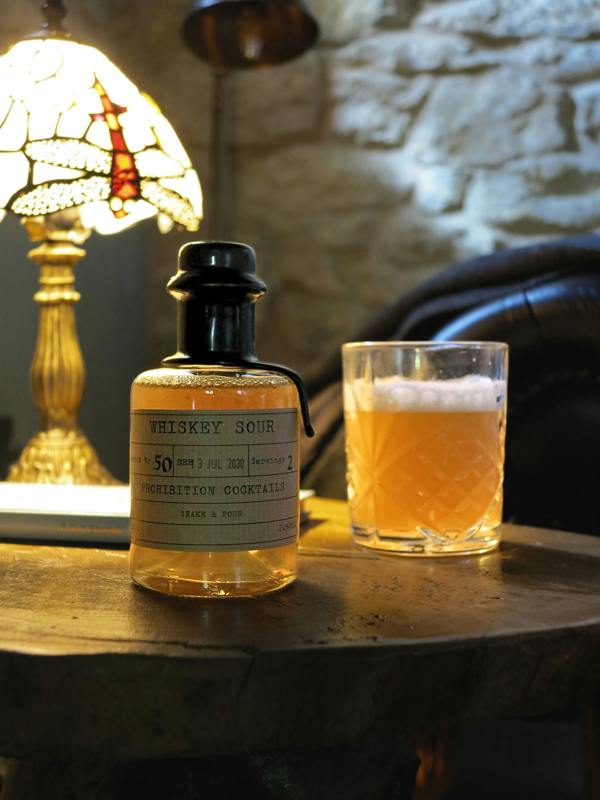 A bottle of Whiskey Sour cocktail mix and a glass of the prepared amber drink sit on a rustic wooden table in a warm, dimly lit setting with a stained-glass lamp and stone wall in the background.