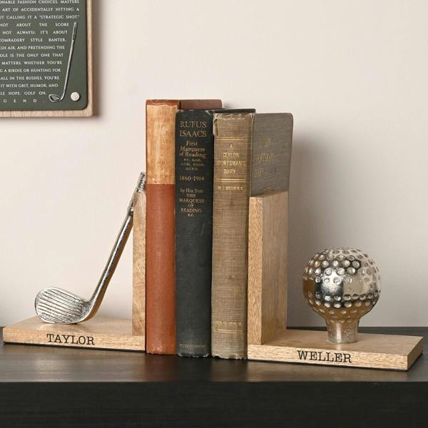 A pair of personalised wooden golf bookends holding three vintage books on a dark shelf, featuring an engraved club and a golf ball on a tee with the names 'Taylor' and 'Weller'.