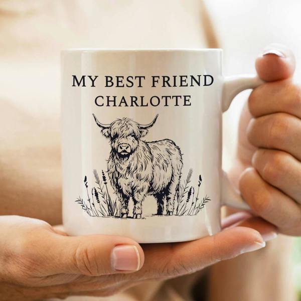 A person holding a white ceramic mug featuring a black illustration of a Highland cow and the text 'MY BEST FRIEND CHARLOTTE' in uppercase.