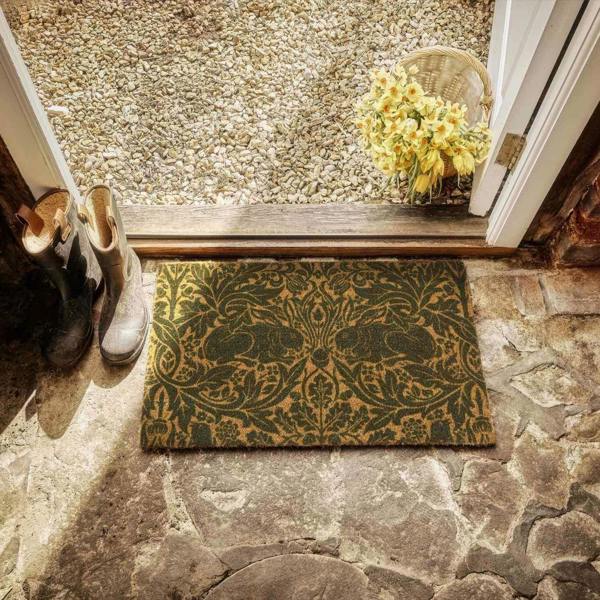 Decorative doormat with floral design on a stone floor, with shoes and flowers in the background.
