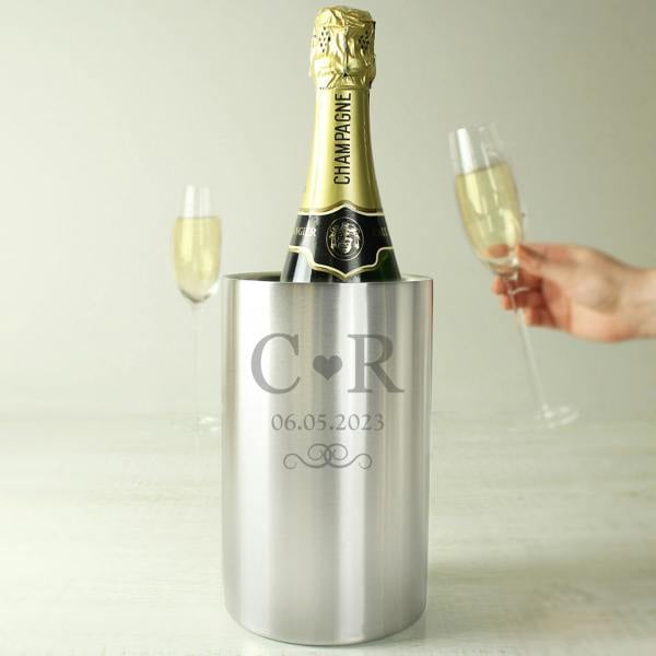 A brushed stainless steel wine cooler engraved with initials C and R separated by a heart, holding a champagne bottle next to two filled flutes.