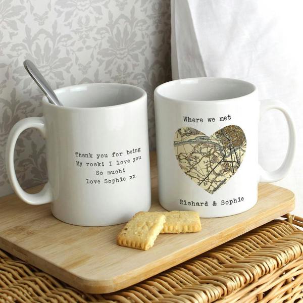 Two custom ceramic mugs from Abode Home & Gifts on a wooden tray, one showing a heart-shaped postcode map and the other showing an engraved love note.
