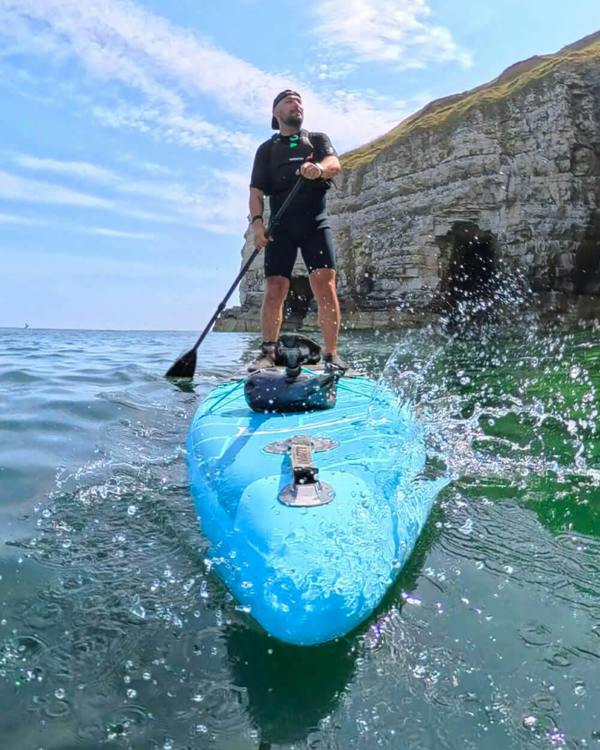 Person paddleboarding on a bright blue SUP in calm waters near a rocky shoreline on a sunny day.