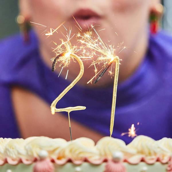Lit gold number 21 sparkler cake toppers sparkling brightly on top of a frosted birthday cake, with a person blowing them out in a blurred background.