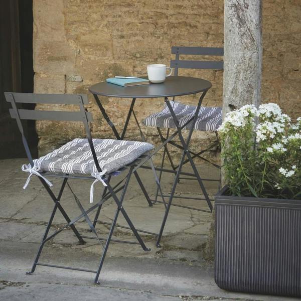 Carbon grey metal folding bistro table and two chairs styled with striped cushions on a stone patio.