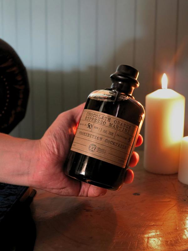 A hand holds a dark glass bottle labeled 'Chocolate Orange Espresso Martini' and 'Prohibition Cocktails' in front of a warm, candlelit background with a lit pillar candle.