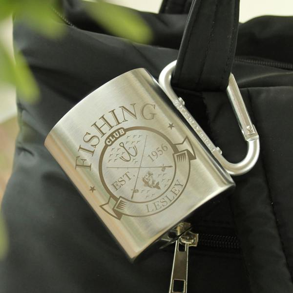 Personalised stainless steel camping mug with a carabiner handle engraved with a "Fishing Club Est. 1956 Lesley" badge, clipped to a black rucksack.