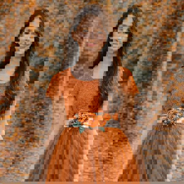 Young girl in an orange dress with a floral sash standing in an autumn setting.