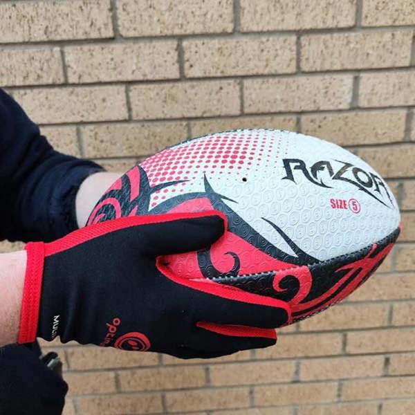Hands in black and red Optimum Multi-X gloves holding a white and red Razor rugby ball on brick - Optimum