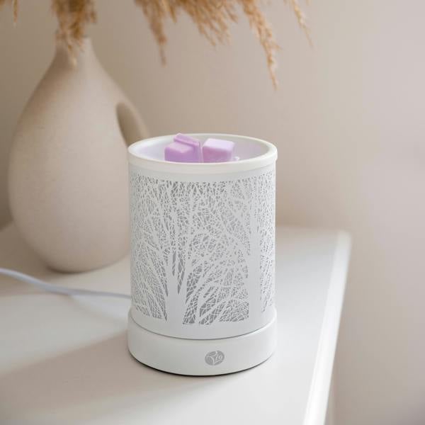 The Rio FORA Wax Melt Burner & Aroma Diffuser Lamp, featuring a tree branch pattern, melts two purple wax cubes on a white surface. A beige vase with dried grass in the blurred background adds to the relaxing ambiance.