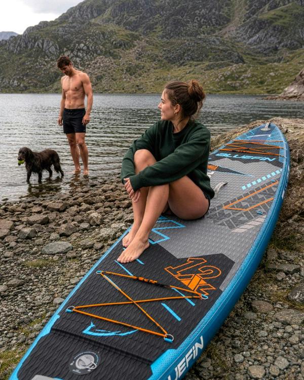 Inflatable paddleboard on rocky shore, woman in green sweater sitting, man with dog in water.
