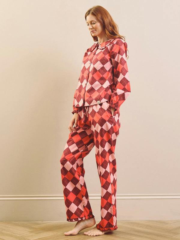 A woman modeling a two-piece pyjama set with a red, pink, and white harlequin diamond pattern, standing barefoot on a light wood floor against a beige wall.