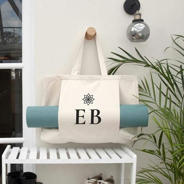 A cream organic cotton yoga tote bag printed with the initials EB under a mandala emblem, holding a green mat.